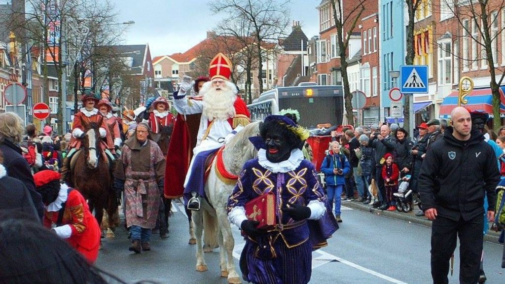 Sinterklaas wird in der nordniederländischen Stadt Groningen empfangen. Dem Bischofsdarsteller zu Pferd geht ein traditioneller Zwarte Piet mit schwarzer Gesichtsfarbe voran. Nach einem Zug durch die Stadt trifft Sinterklaas den Bürgermeister. Rechts im Bild sieht man einen Wachmann. - Bild: Berkh, CC BY-SA 4.0, via Wikimedia Commons